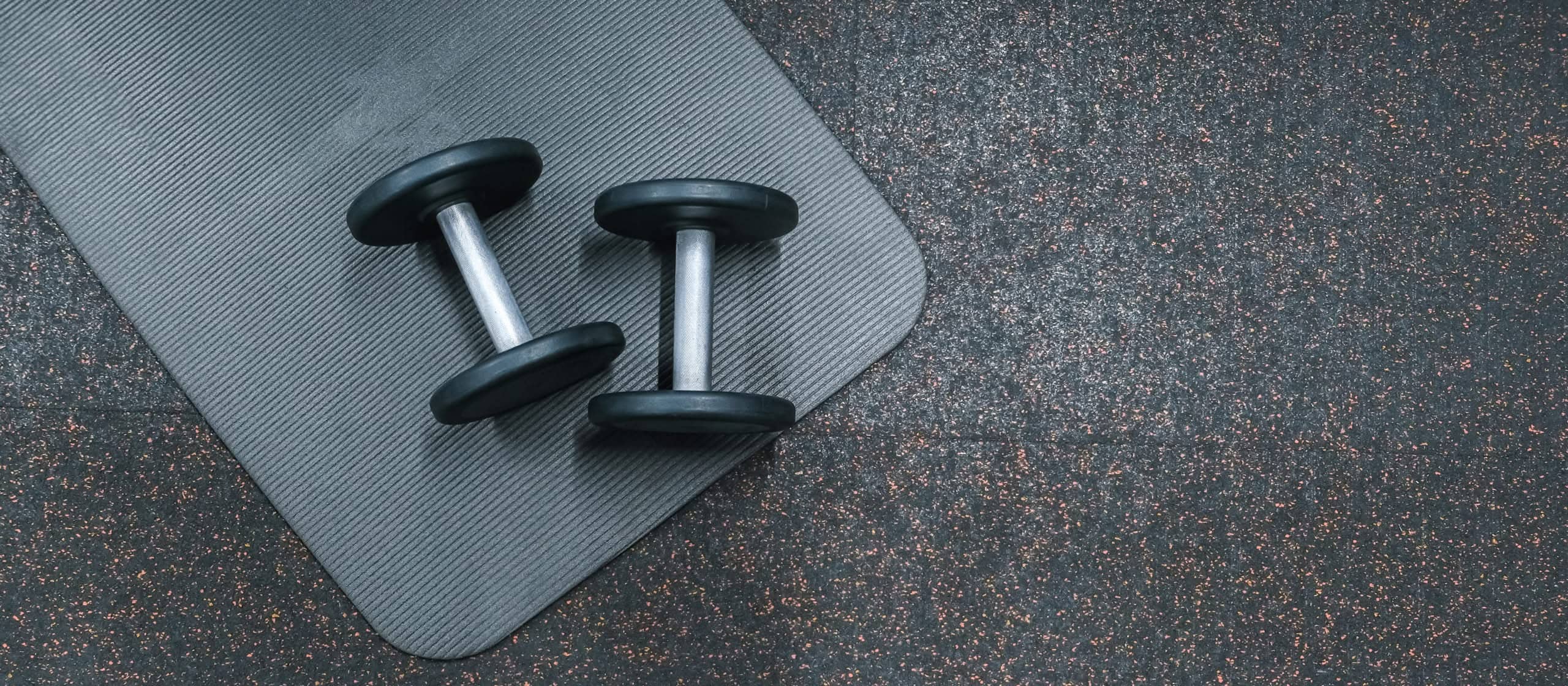 Top-down view of two black dumbbells sitting on a ribbed grey exercise mat laid over speckled rubber gym flooring in Ocala, FL.